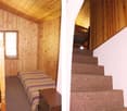 The image shows the interior of a wooden cabin. The walls and ceiling are made of wood panels, creating a cozy and rustic atmosphere. The room has a bed with striped bedding, and there is a staircase leading to another level. The overall impression is of