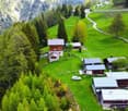 Aerial view of a rural mountain village surrounded by lush green forests and meadows. The village consists of traditional wooden cabins and houses with red roofs. There are paved roads and a few vehicles visible. The landscape is characterized by rolling
