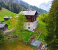 A traditional alpine-style wooden and stone house nestled in a lush, green mountain landscape. The house has a sloped roof, balconies, and is surrounded by tall pine trees and rolling hills.