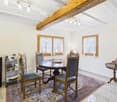 Spacious dining room with wooden beams on the ceiling, a round wooden dining table with chairs, a rug on the floor, and a fireplace visible in the room.