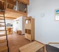 Open-concept studio apartment with wooden beams, loft area, kitchen, dining table, shelving unit, and a framed artwork on the wall.