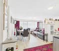 Open-concept living room and dining area, hardwood floors, large windows, purple curtains, leather sofa, dining table with chairs, shelving unit with decor, rug