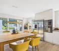 Spacious open-plan kitchen and dining area with large windows, wooden dining table and yellow chairs, built-in kitchen appliances including a refrigerator and oven, and decorative accents.