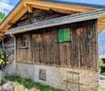 Rustic wooden cabin with a balcony, stone foundation, and a green window. The cabin is surrounded by lush greenery and mountains in the background.