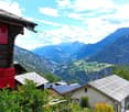 Picturesque mountain village with traditional wooden houses, red shutters, and solar panels on the roofs. The village is surrounded by lush green mountains and a scenic valley in the background.