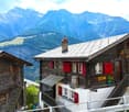 Rustic wooden mountain cabins with red shutters, set against a backdrop of snow-capped mountains. The cabins have balconies and appear to be located in a scenic alpine setting.