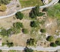 Plot of land with a small house, with green grass and trees, visible from above, surrounded by a road and other properties.