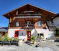 Brown wooden building, 2 balconies, red entrance door, awnings, barrels, plants