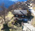 House with a raised ground floor, wooden exterior, grey roof, white walls, garage, located on a mountain road, driveway leading to the house, trees, mountainous surroundings