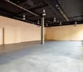 Large open commercial space with concrete flooring, exposed ceiling beams and ductwork, and track lighting. The space appears to be a studio or showroom with a fire extinguisher visible in the corner.