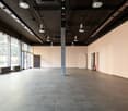 Large open commercial space with concrete flooring, exposed ceiling with lighting fixtures, and large windows along one wall.
