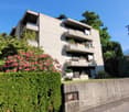 3-story apartment building with balconies, surrounded by lush greenery and trees, with a concrete retaining wall in the foreground