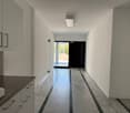 The image shows a long, narrow hallway with white walls and a tiled floor. The hallway has built-in cabinets or storage units along one side, and a doorway at the end leading to an outdoor area. The lighting appears to be recessed and the overall aestheti