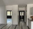 The image shows the interior of an apartment. The space features a modern, minimalist design with white walls and cabinets, and gray tile flooring. There is a kitchen area with a granite countertop and a dishwasher visible. The apartment has large windows