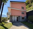 Pink house, gray brick base, concrete driveway, balcony on the second floor, front yard with lawn, bushes, palm tree, mountains in the background