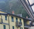Two-story yellow building with several balconies, green shutters, and a chimney. Set against a backdrop of a hillside with trees.
