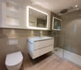 Modern bathroom with white vanity, large mirror with lighting, tiled walls and floor, shower enclosure, and toilet