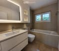 Spacious bathroom with a bathtub, vanity sink, and storage cabinets. The room has tiled floors and walls, and a window overlooking the outdoor greenery.