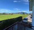 Apartment balcony with metal railing, paved area for car to park, view of mountains and a field