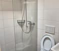 A compact bathroom with a glass shower door, white tiles, a silver shower head, a toilet, and a sink
