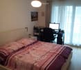 Large bed, pink blanket, two pillows, desk, monitor, chair, small white shelf, picture, curtains