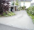 Driveway leading to garage with car parked inside, surrounded by greenery, two manholes, house with a terrace in the background.
