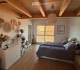 Bedroom with wooden floor, white walls, wooden ceiling beams, large window, bed, nightstand, cabinet, guitar, and decorative items.