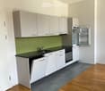 Modern kitchen with white cabinets, black countertops, and stainless steel appliances including a refrigerator, oven, and dishwasher. Green tiled backsplash and wooden flooring.