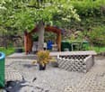 A garden with a wooden gazebo, a table and chairs, a potted plant, a blue barrel, a green tank, a yellow hose, and a stone wall.