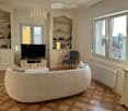 Living room with a large curved sofa, a flat screen TV, a wooden entertainment center, a blue chair, a white radiator, and large windows with a view of the outside.