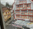 Aerial view of a building with multiple floors, red trim, and many windows. The building has a name board that reads 'BAZAR HERSCHE'. The building has a small garden with a few potted plants. The building is surrounded by other buildings and a street.