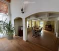 Bright hallway with wooden flooring, large potted plant, arched entryway leading to a dining area with a wooden table and chairs, visible kitchen area with cabinets and a clock on the wall.