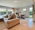 Spacious living room with beige sofa, coffee table, dining table, and chairs. Large windows with curtains. Beige rug and decorative items.