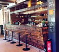 Wooden bar counter with hanging wine glasses, bottles, and stools, with a fire extinguisher nearby.