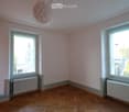 Empty room, white walls, white ceiling, white radiators, two windows, wooden parquet flooring, view of stairs from one window
