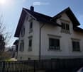 House. Two-story house with a brown roof, white walls, and green shutters on the windows. The house is surrounded by a wooden fence.
