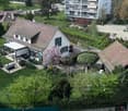 Aerial view of a residential area with a single-family house, garden, patio, and surrounding greenery. The house has a tiled roof, multiple windows, and a covered outdoor seating area. The garden includes lawn, trees, and shrubs.