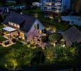 A modern two-story house with a tiled roof, surrounded by a well-maintained garden and illuminated pathways. The house features large windows and a covered outdoor seating area with a table and chairs. Nearby, there are additional residential buildings an
