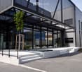 Building. Modern building with large glass windows and a sleek, contemporary design. Features a raised ground floor, small water feature, and small landscaped area with plants and trees.