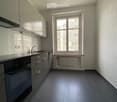 Modern kitchen with gray cabinets, black appliances, and a large window providing natural light. The kitchen has a tiled floor and appears to be in a residential apartment or condominium.
