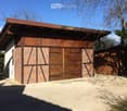 Building. A wooden garage with a brick facade and large wooden doors, situated on a gravel driveway under a clear blue sky.