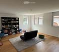 Modern living room with wooden flooring, large windows, a dark grey sofa, a blue armchair, a bookshelf filled with books, and a small rug.