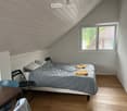 Cozy attic bedroom with wooden slat ceiling, grey bedding, two chairs, and a small table.
