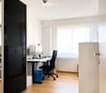 Modern office space with wooden flooring, large windows, a desk with dual monitors, a black office chair, and a tall cabinet.