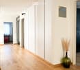 A modern hallway with light wooden flooring, white walls, built-in closet, and a decorative vase with dried plants on the floor.