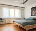 A modern bedroom with a wooden bed, grey duvet, a black chair, a radiator, a ceiling fan, and a colorful abstract painting on the wall.