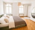 Bright bedroom with a large bed, green sofa, white pillows, a round mirror, a wardrobe, and a pendant light hanging from the ceiling.