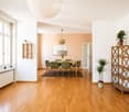Bright, open-plan living and dining area with wooden flooring, large windows, and modern furniture including a dining table, chairs, shelves, and potted plants.