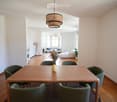 Bright, open living and dining area with wooden flooring, large windows, a dining table with six chairs, a sofa, an ottoman, a floor lamp, and decorative vases.