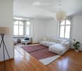 Bright living room with white walls, wooden flooring, large arched windows, a white sectional sofa with ottoman, two white armchairs, a small side table, a floor lamp, and a potted plant.